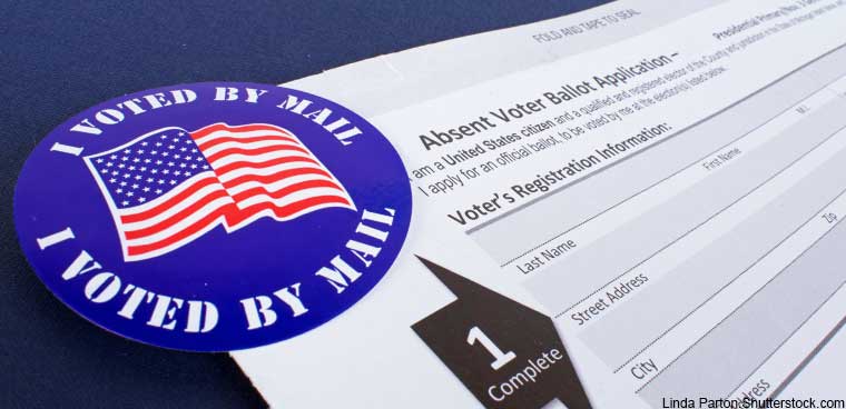 An+image+of+a+blue+I+voted+by+mail+sticker%2C+with+an+American+flag+displayed+over+it.+Beneath+the+sticker+is+a+blank+white+voting+ballot.
